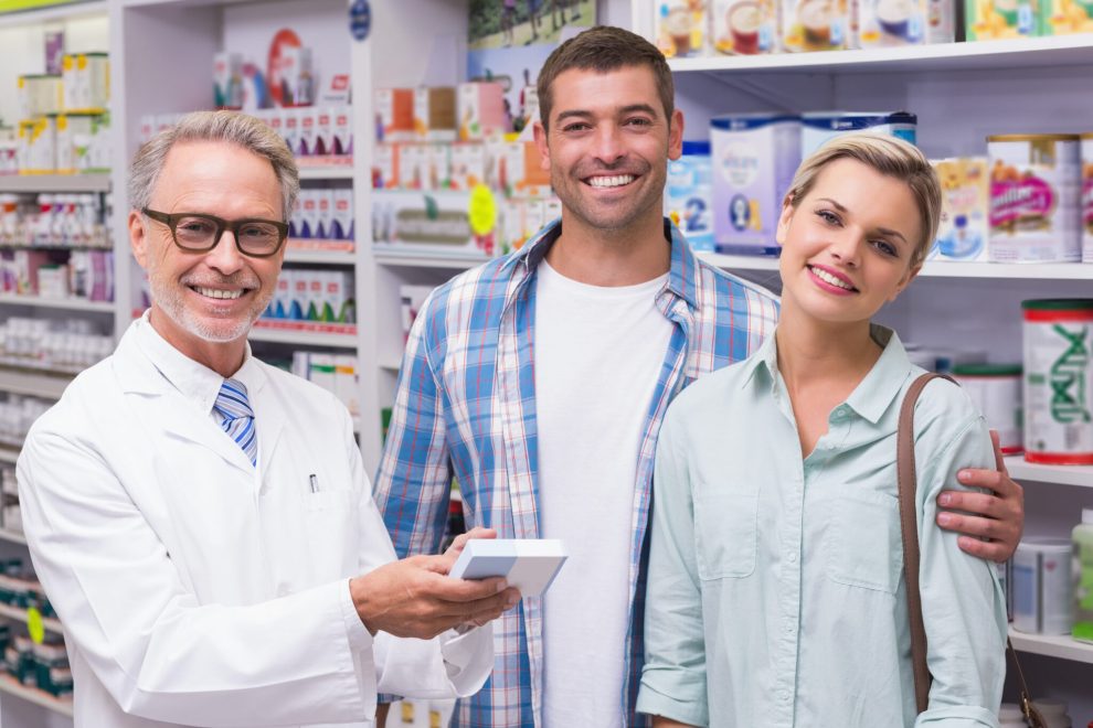 Pharmacien souriant avec clients, pharmacie en arrière-plan.